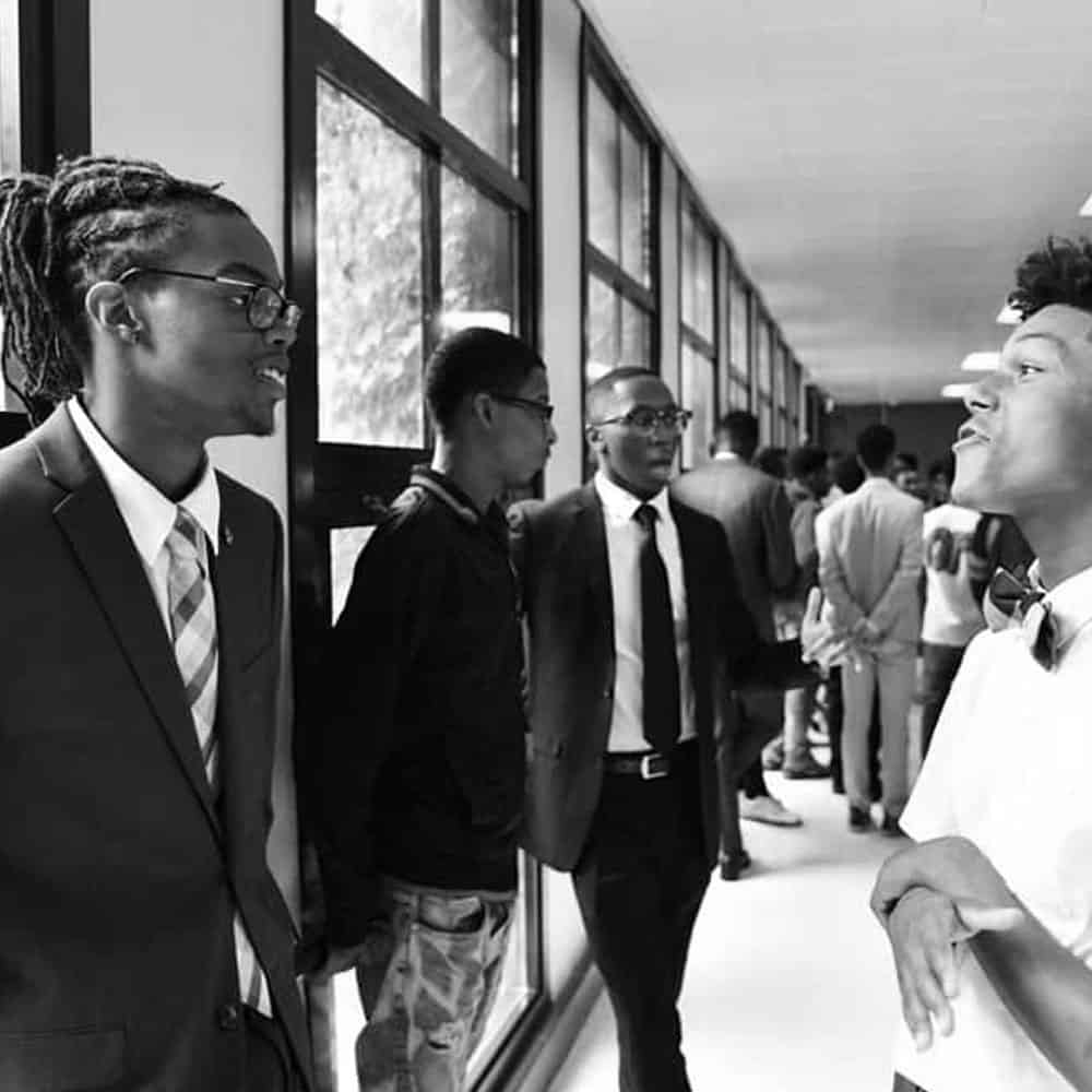 We Are Confidence: A group of young men, dressed in suits and casual attire, stand and converse in a hallway with large windows.