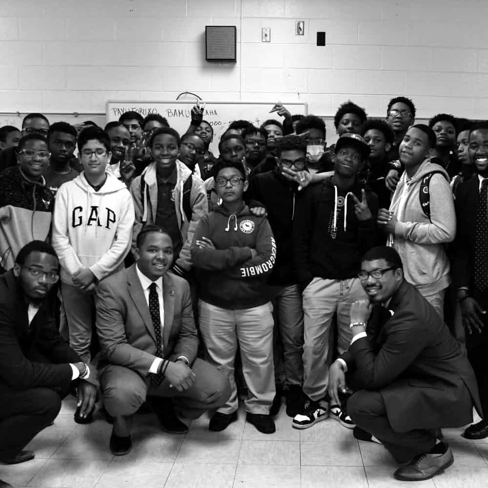 We Are Excellence: Group of young men and several adults in suits posing together in a classroom, some smiling and showing peace signs.