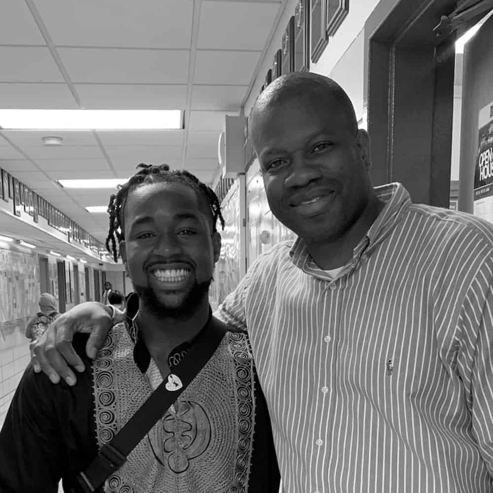 We Are Respect: Two people standing side by side, smiling in a hallway. The person on the left is wearing a patterned shirt, and the person on the right is wearing a striped shirt.