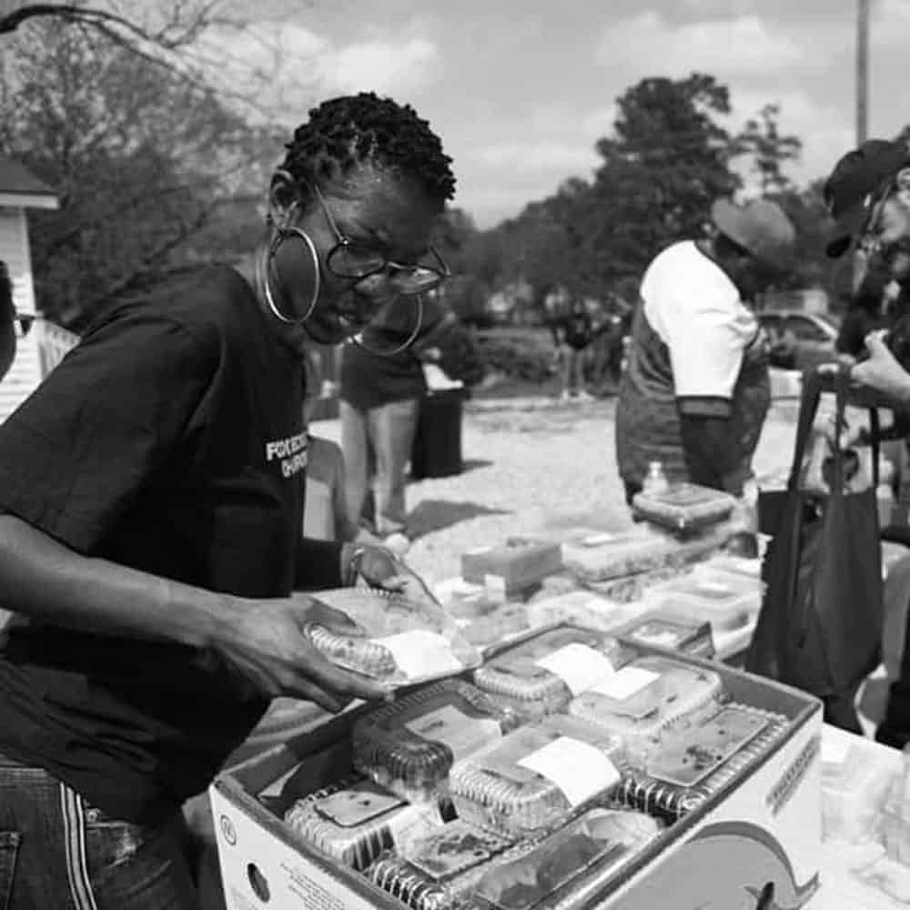 We Are Servant Leadership: A person in a t-shirt sorts food packages outdoors, while others gather nearby. There are boxes filled with food items on a table.