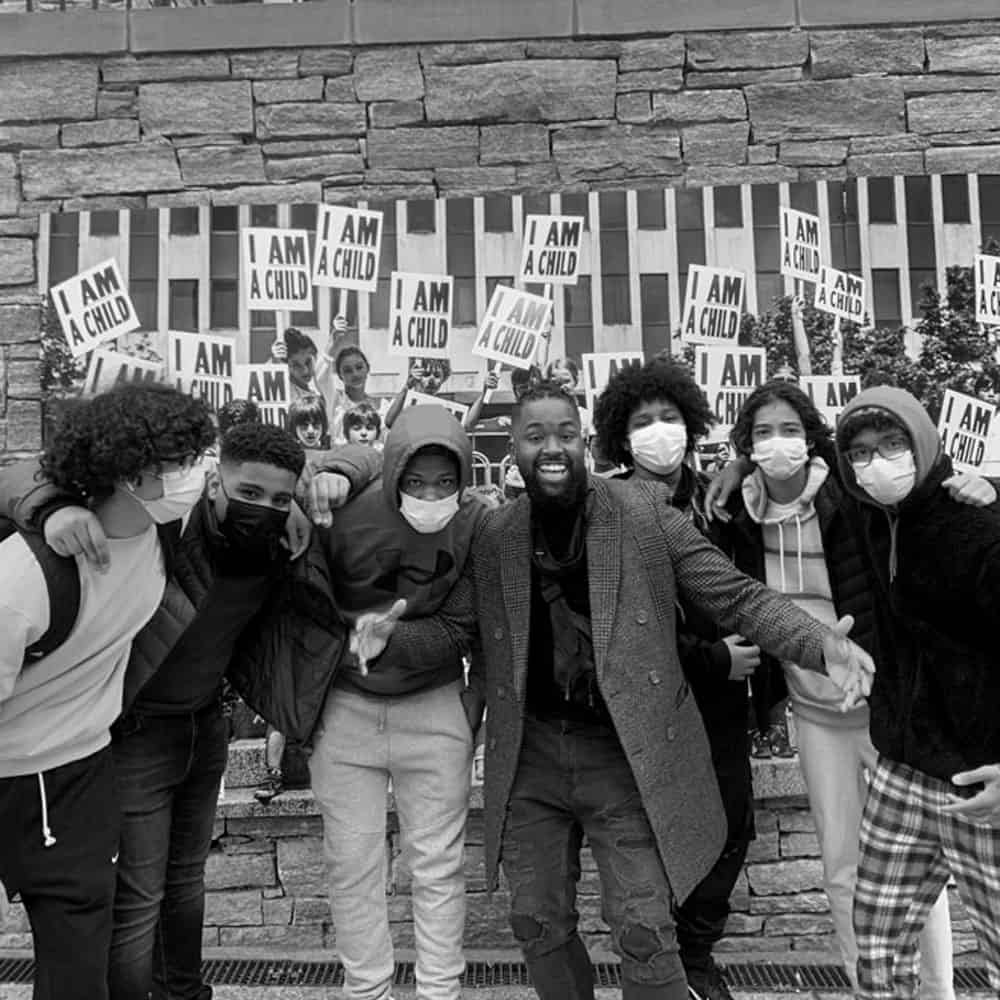 We Are Community: A group of people pose in front of a brick wall, that has a mural of children holding signs that read "I AM A CHILD."